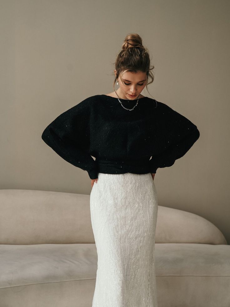 a woman standing in front of a couch wearing a black sweater and white skirt