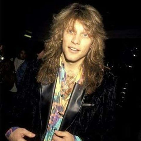 a man with long hair wearing a leather jacket and colorful neck tie standing in a dark room