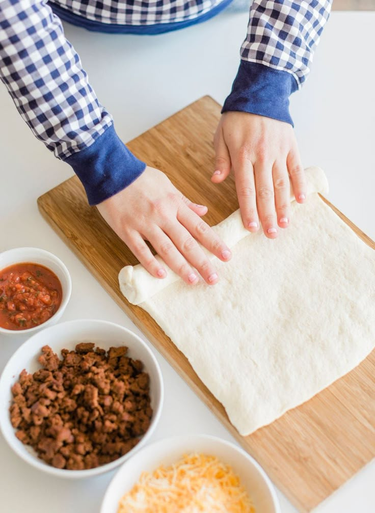 a person is kneading dough on a cutting board next to bowls of chili and cheese