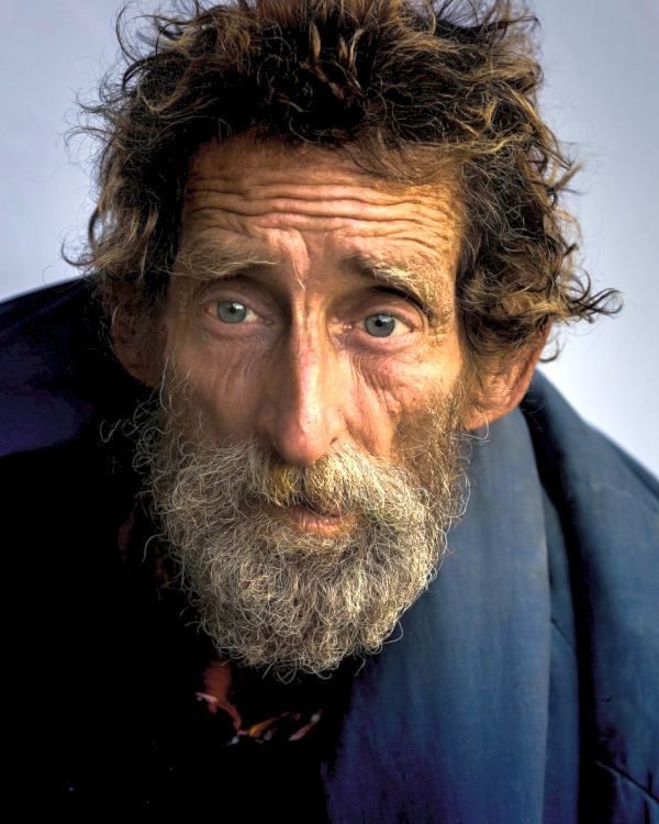 an old man with long hair and a beard looks at the camera while wearing a blue jacket