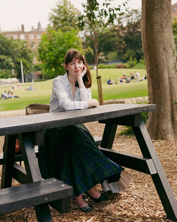 a woman sitting at a picnic table with her chin resting on her hand, in a park