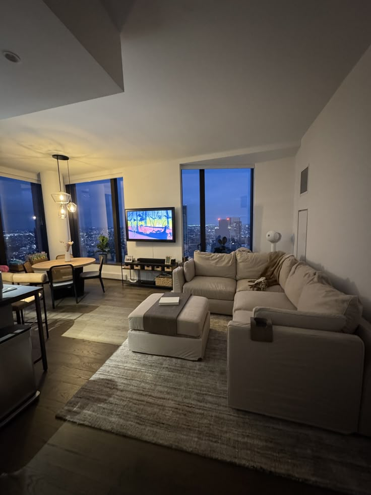 a living room filled with furniture and large windows overlooking the city at night, in front of a flat screen tv