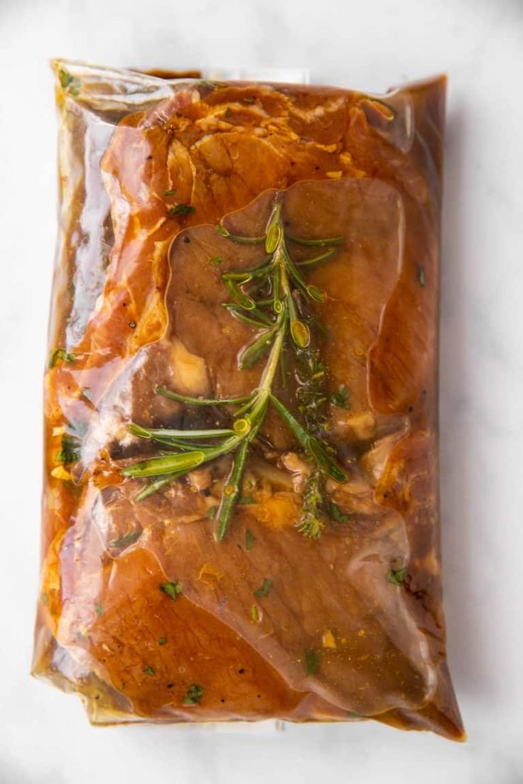 meat wrapped in plastic sitting on top of a white counter next to vegetables and herbs