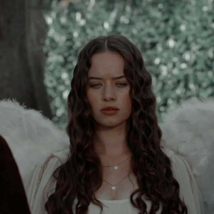 a woman with long curly hair standing in front of an angel's wing and looking at the camera