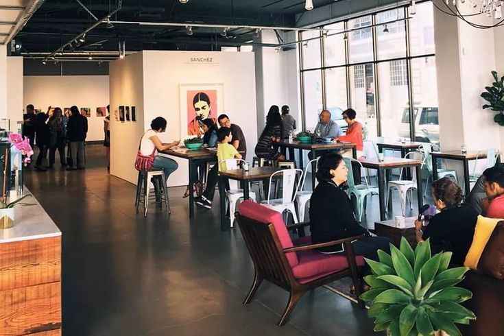 people are sitting at tables in an open space with plants and paintings on the walls