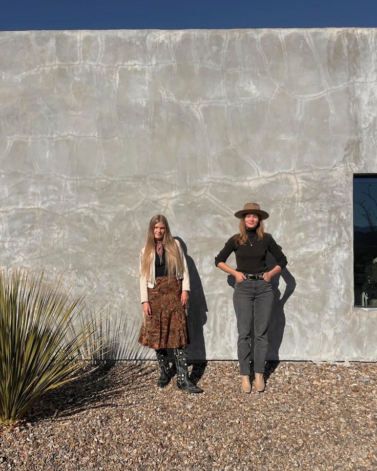 two women standing next to each other in front of a white wall and cactus plant