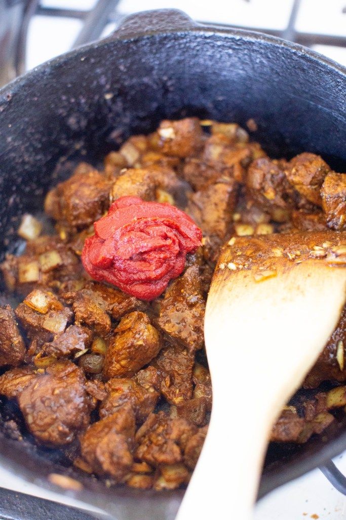 a pan filled with meat and vegetables on top of a stove next to a wooden spoon