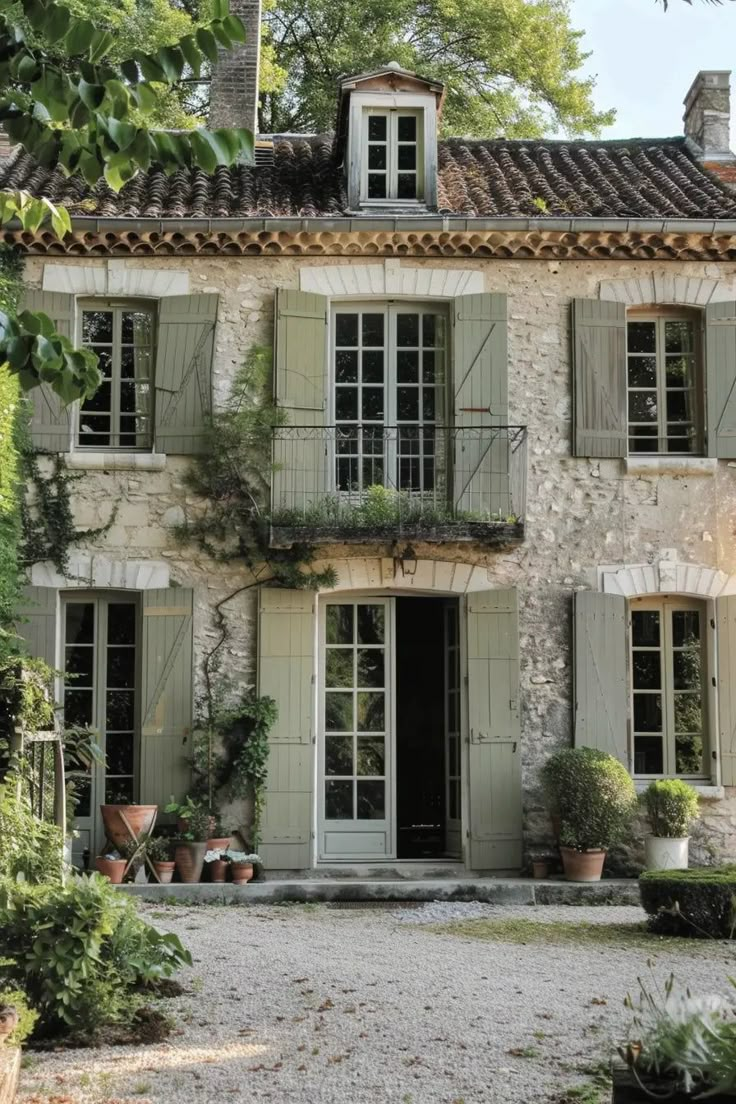 an old stone house with white shutters and plants on the front door, surrounded by greenery