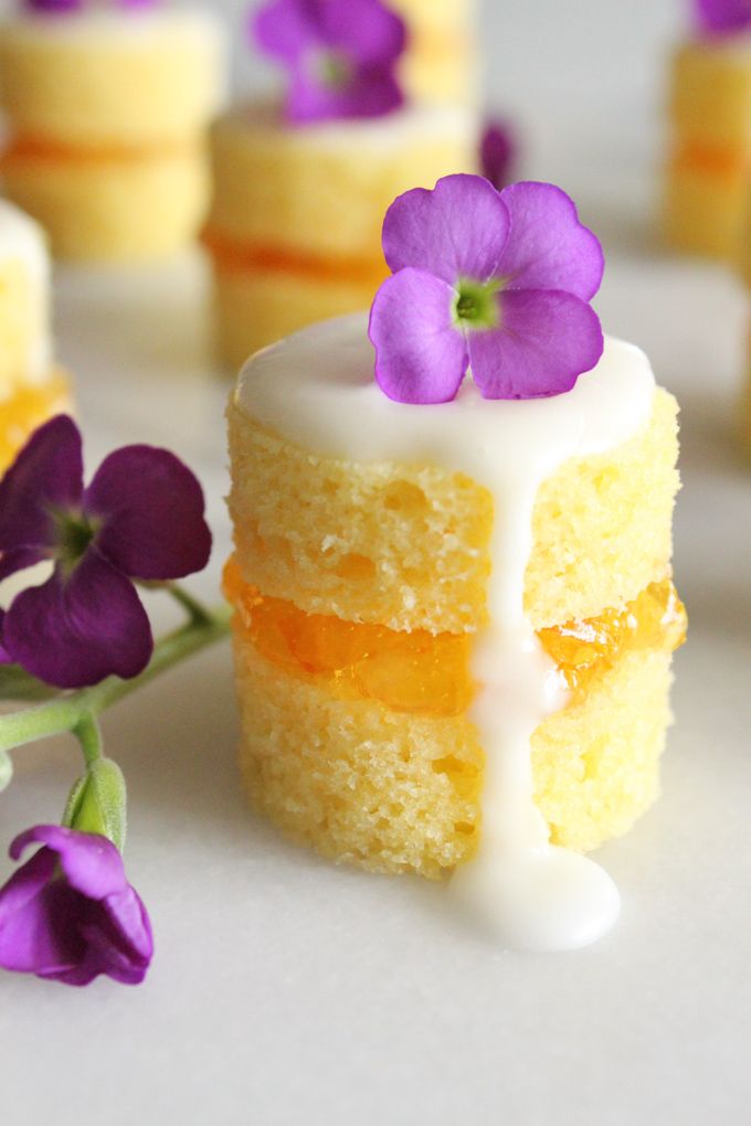 small yellow cake with white icing and purple flowers on it's side, surrounded by mini cupcakes