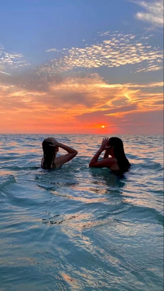 two women in the ocean at sunset with their backs to each other, one laying on her head