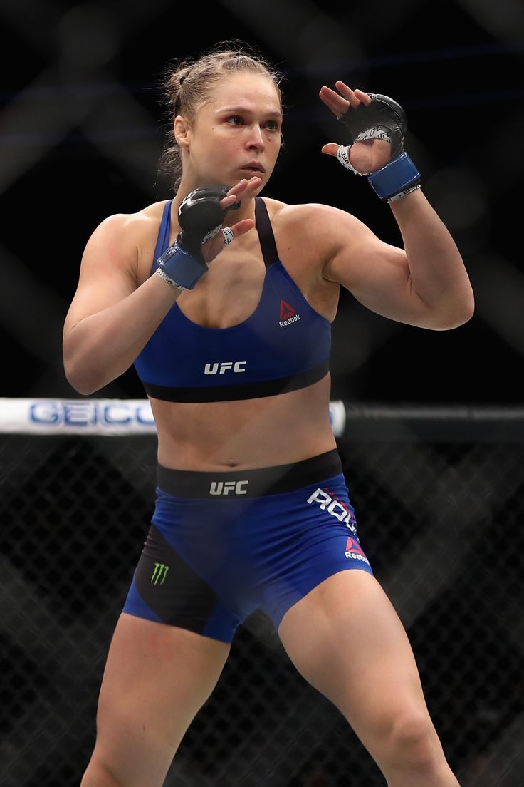 a female fighter in blue shorts and a black top holds her fist up as she prepares to