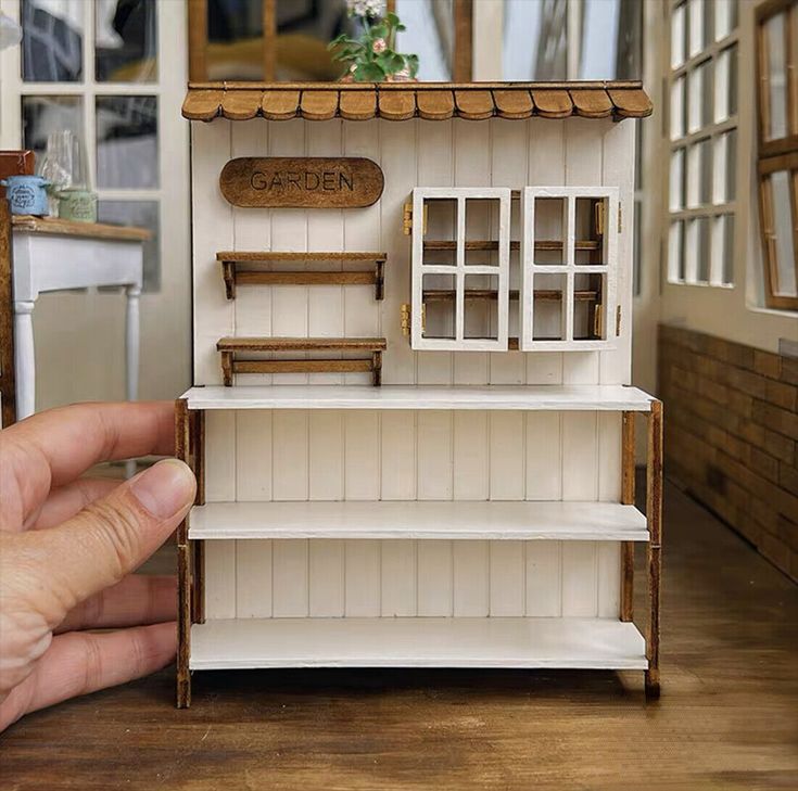 a hand is holding a doll house in front of a window