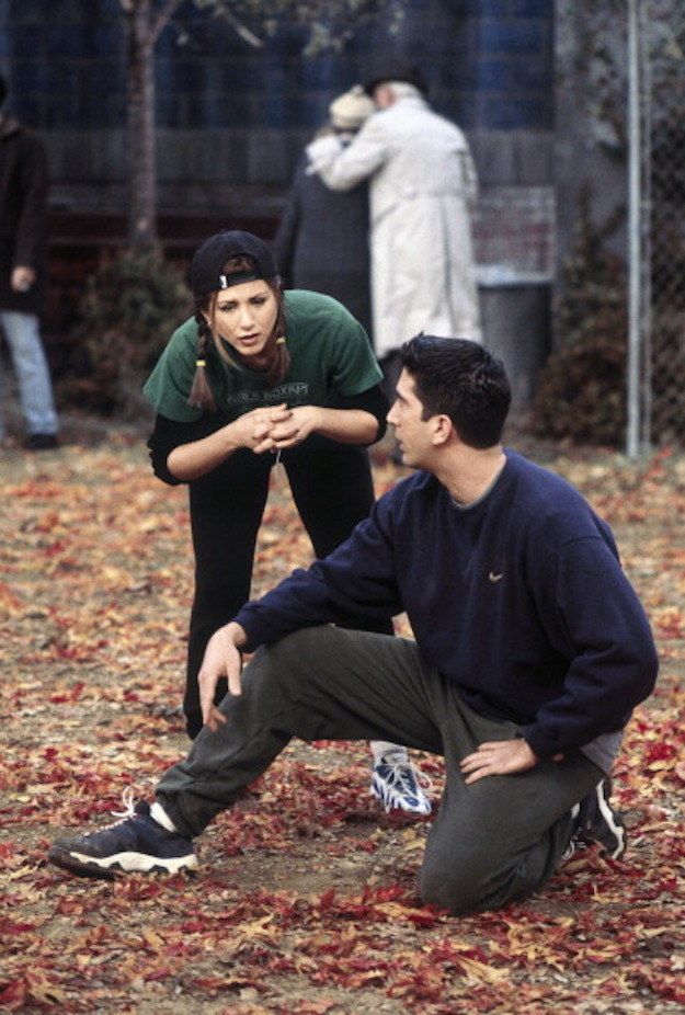 two people sitting on the ground in front of a chain link fence with leaves all around them