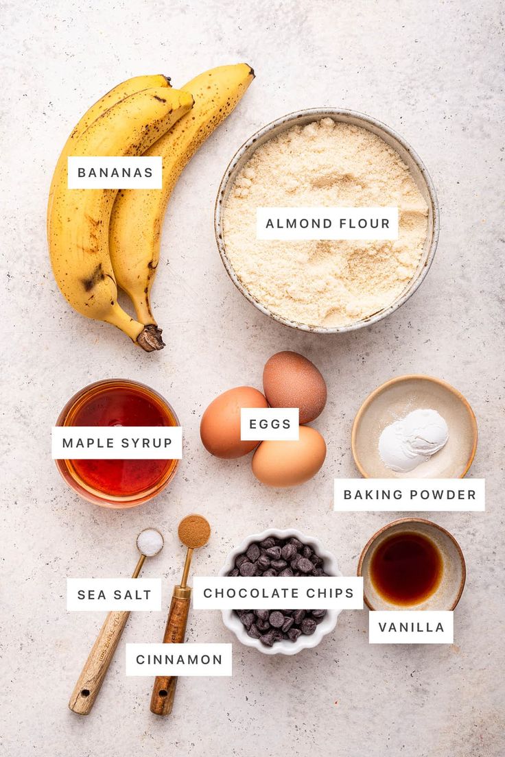 ingredients to make banana bread laid out on a white counter top, including flour, eggs, chocolate chips, and bananas