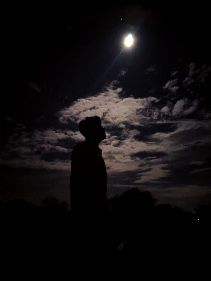 a man standing in the dark with his head turned to the side looking up at the moon