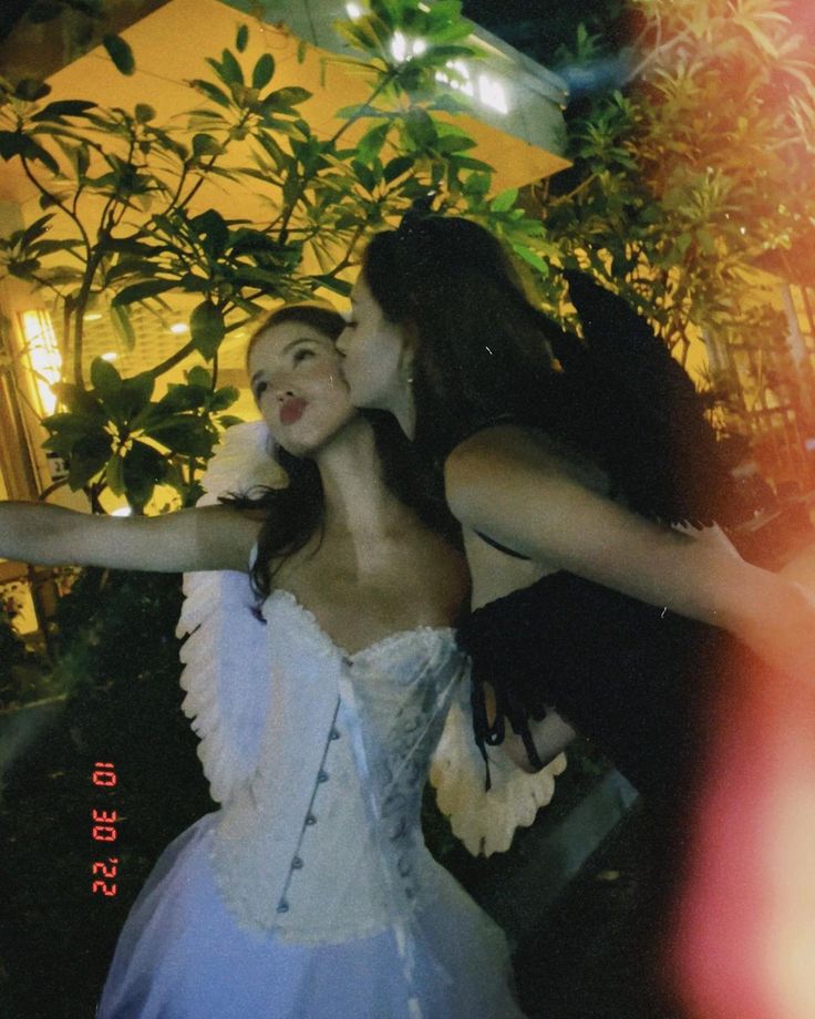 two women dressed in white are kissing each other outside at night with trees and bushes behind them