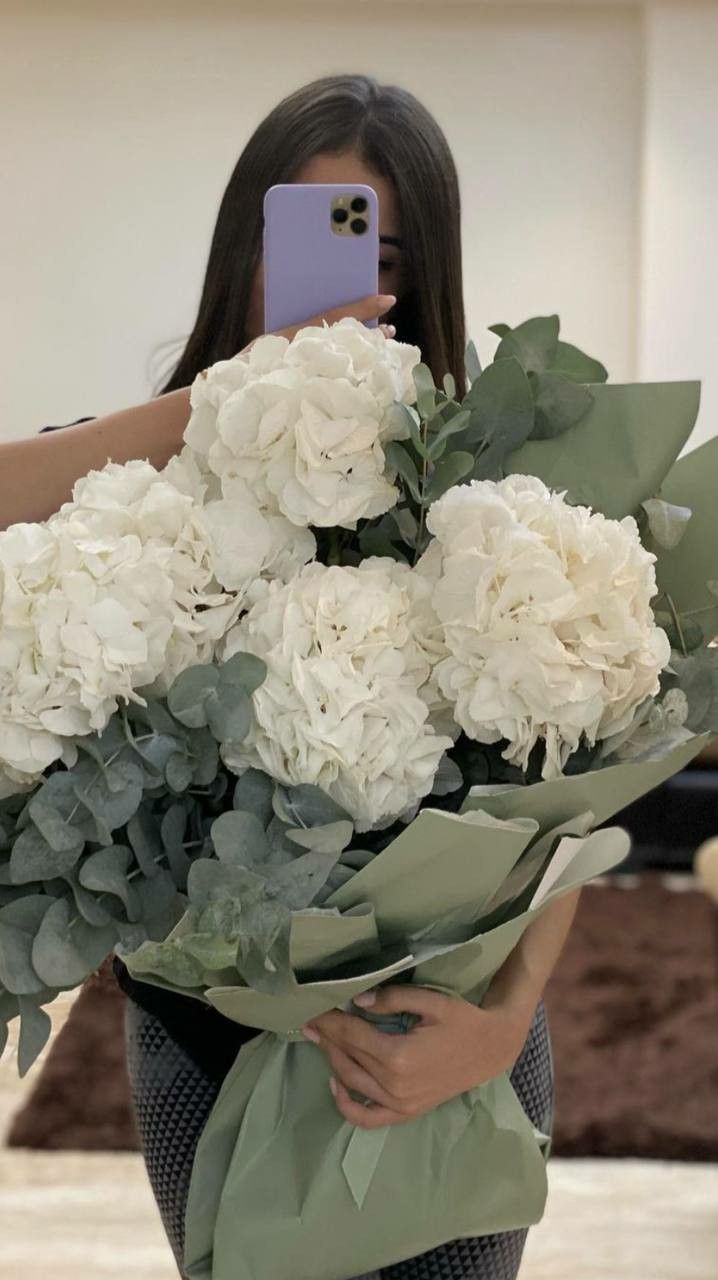 a woman taking a selfie with her cell phone while holding a bouquet of white flowers