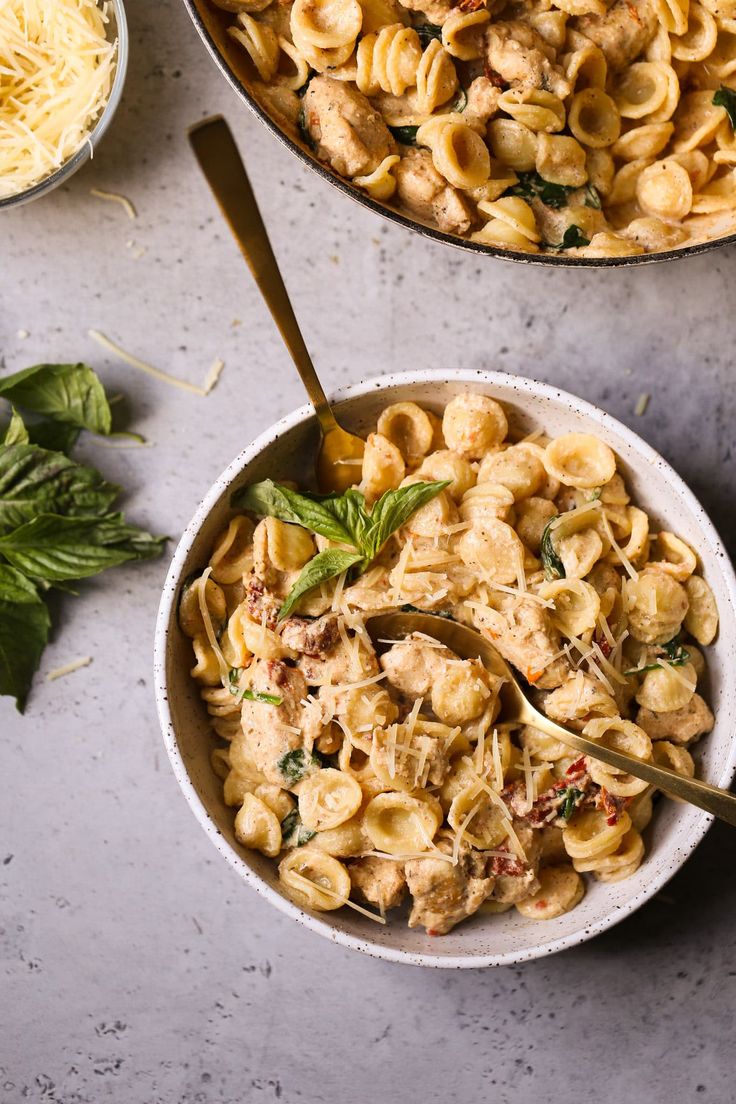 two bowls filled with pasta and chicken on top of a table next to basil leaves