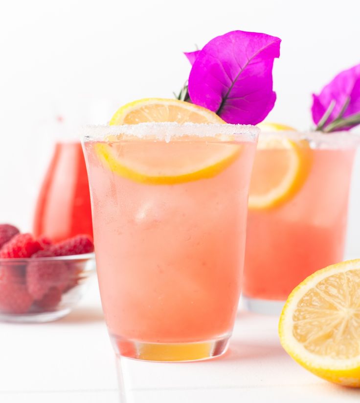 two glasses filled with pink lemonade and raspberries on top of a white table