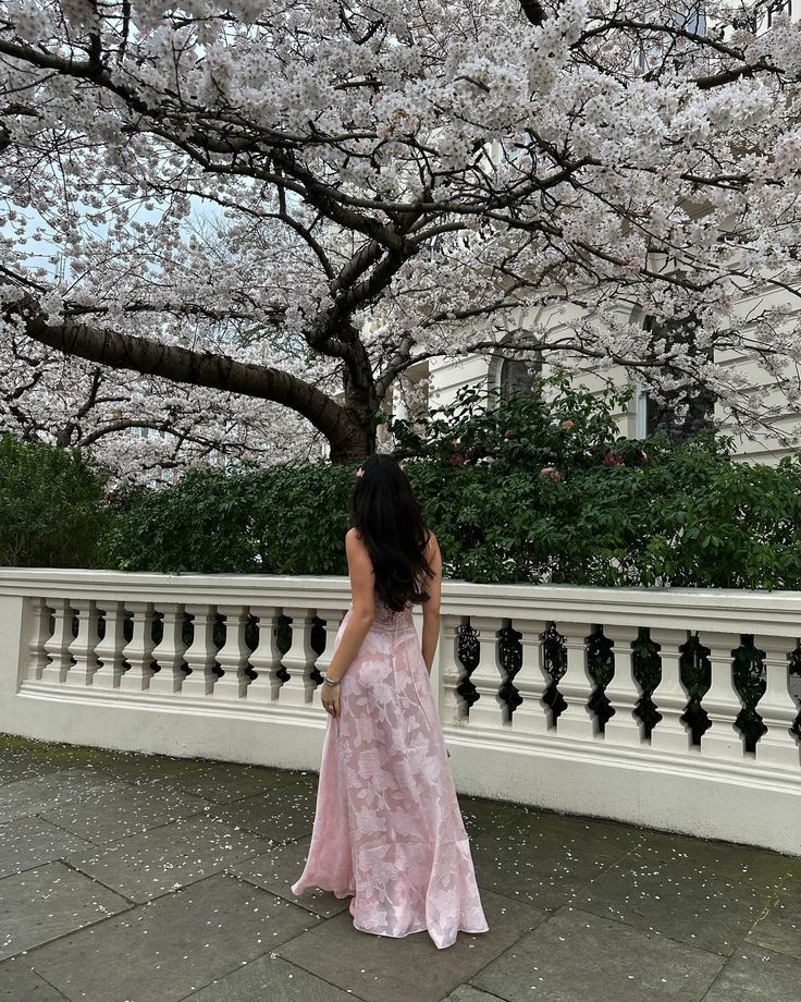 a woman in a long pink dress is standing near a tree with white blossoms on it