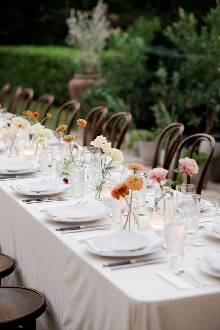 a long table is set with white plates and vases filled with flowers on it