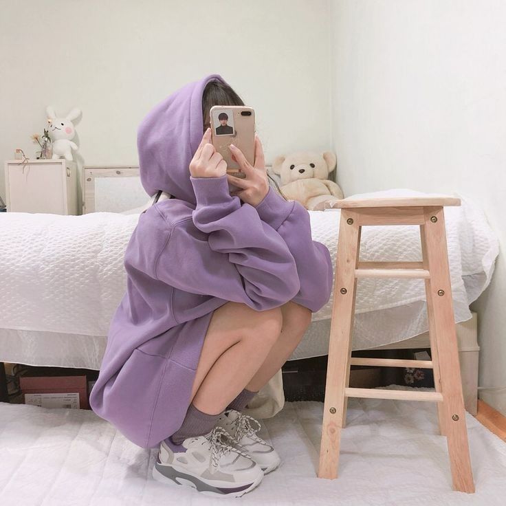 a woman in a purple hoodie taking a selfie with her cell phone while sitting on a bed