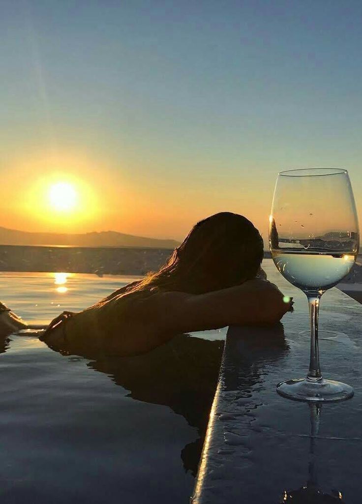 a woman sitting next to a glass of wine on top of a swimming pool at sunset