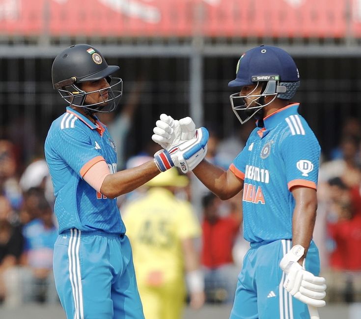 two men in blue uniforms shaking hands at a cricket game with spectators watching from the stands