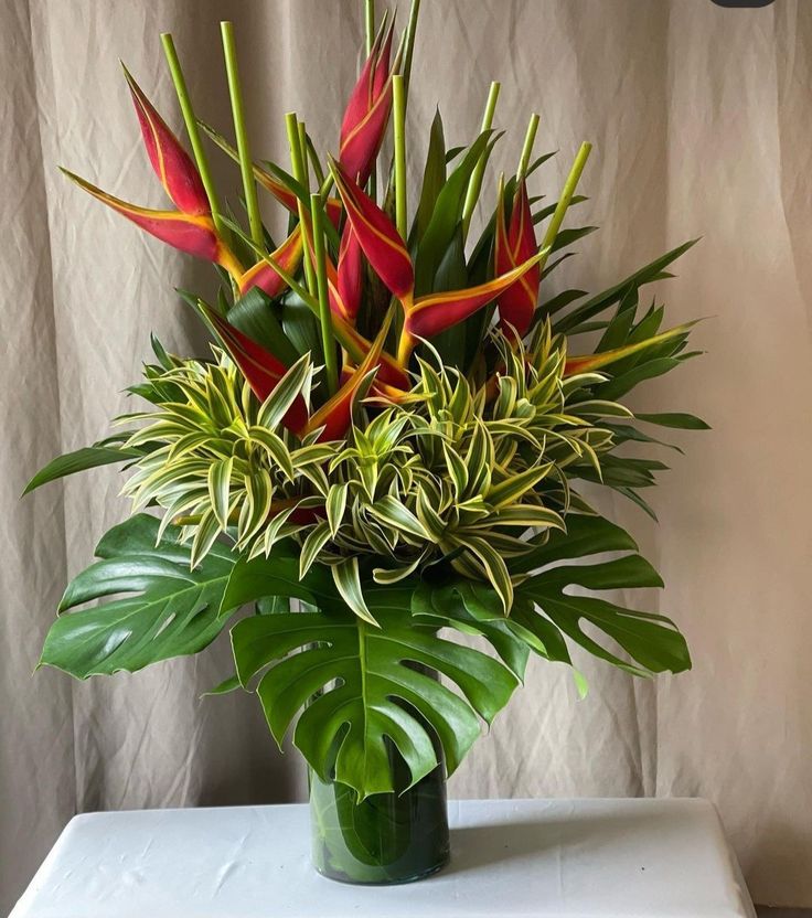 a vase filled with red and green flowers on top of a white table next to a curtain