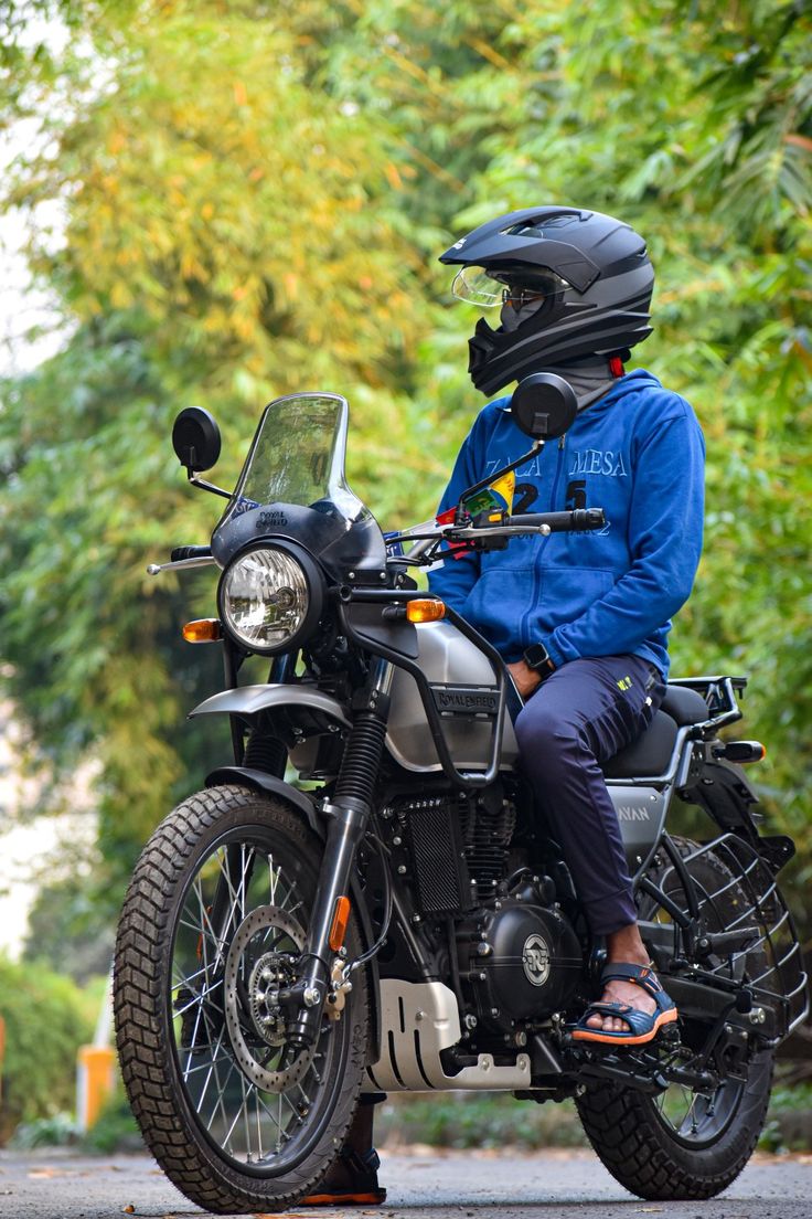 two people sitting on a motorcycle in front of some trees and bushes, with one person wearing a helmet