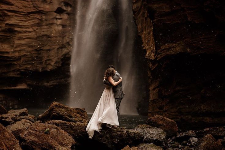 a man and woman standing in front of a waterfall with their arms around each other