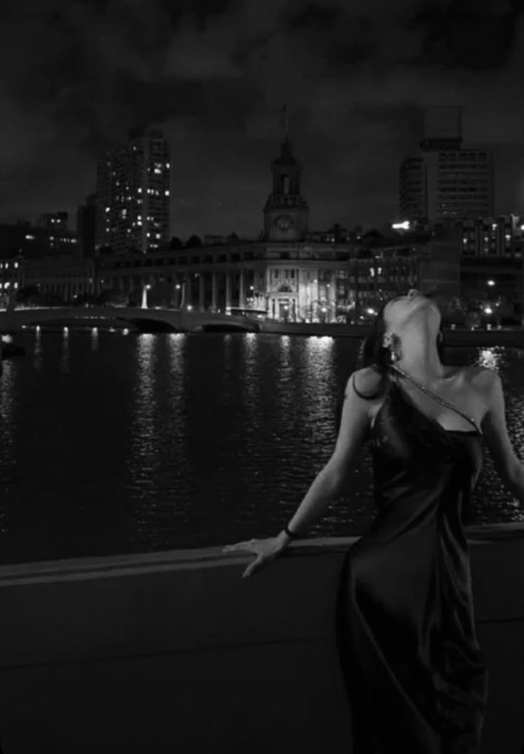 a woman in a long black dress standing on a ledge looking at the city lights