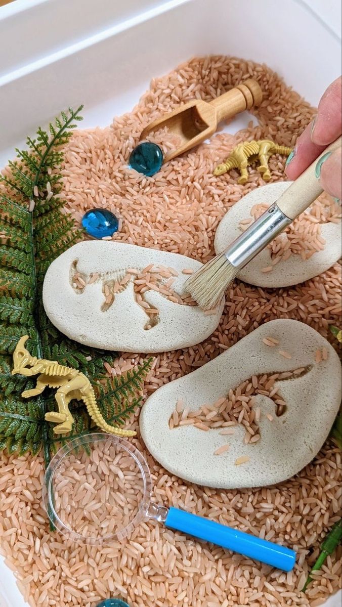 someone is painting fake dinosaur shoes on rice in a box with other toys and materials