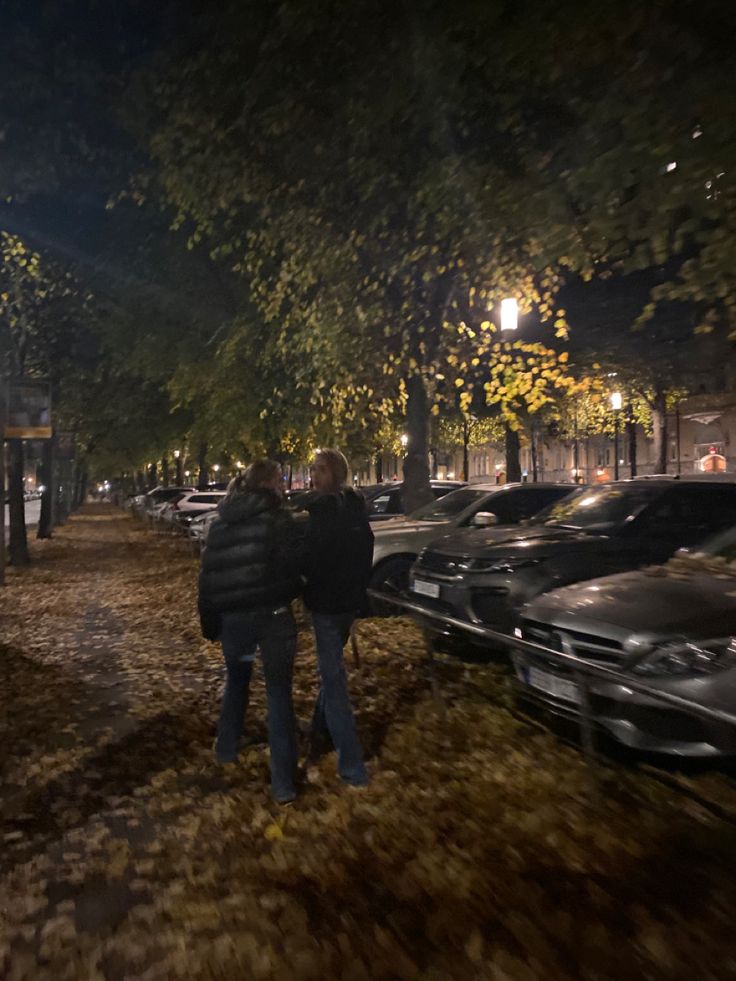 two people walking down the street at night with cars parked on the side of the road