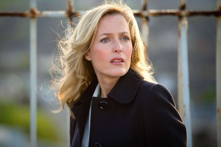 a woman with blonde hair wearing a black coat and looking off into the distance while standing in front of a fence