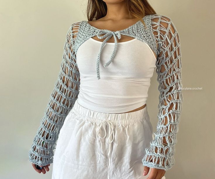 a woman wearing white shorts and a top with crochet on the sleeves is standing in front of a wall