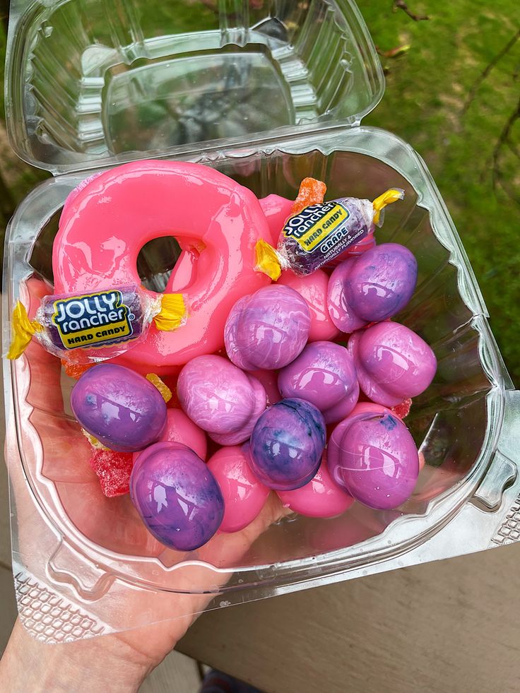 a person holding a pink doughnut with candy on it in a clear plastic container