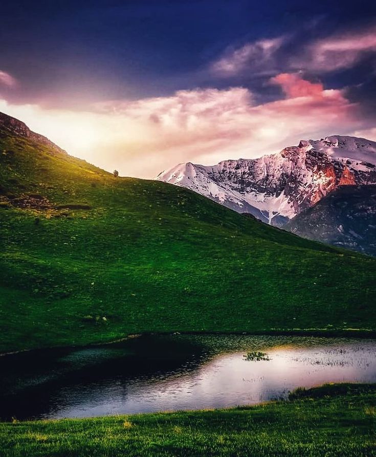 the mountains are covered in snow and green grass as the sun sets over a lake