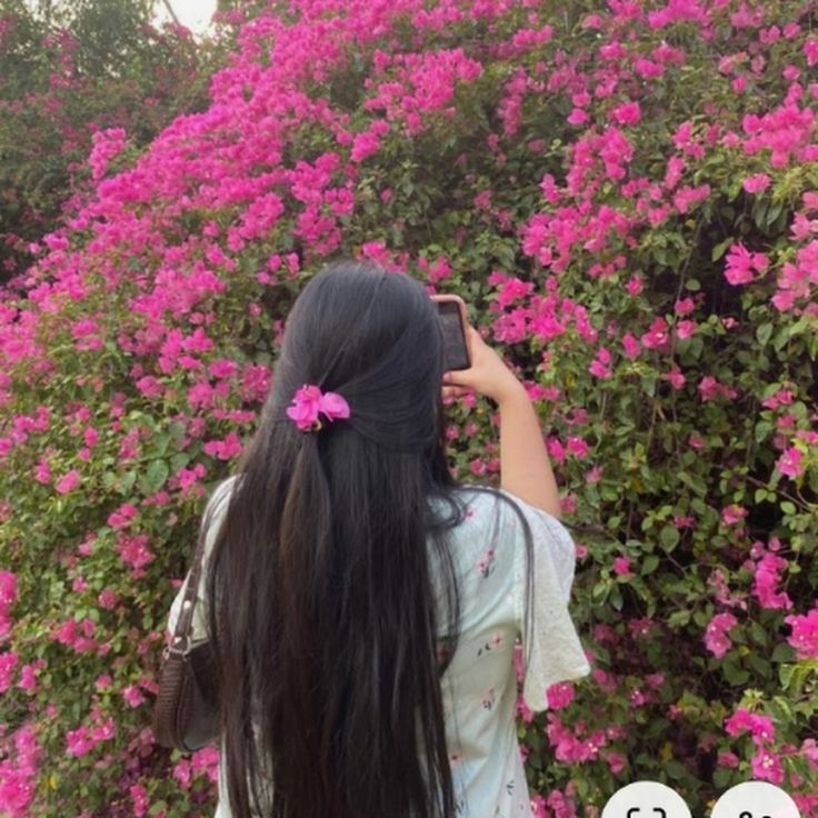 a woman with long black hair standing in front of pink flowers and taking a photo