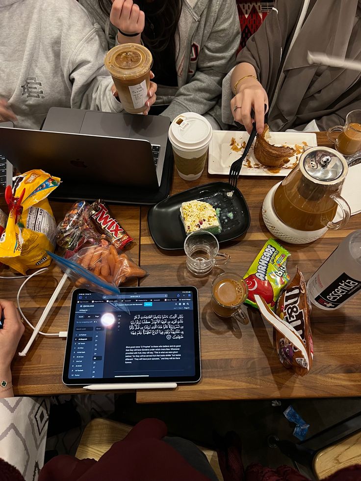 two people sitting at a table with food and drinks in front of their laptops