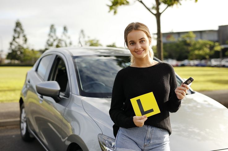 a woman standing next to a parked car holding a yellow sticker with the letter l on it