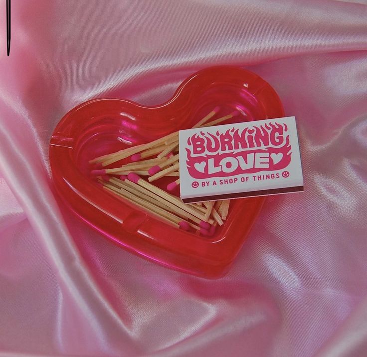 a red heart shaped box filled with wooden clothes pins sitting on a pink satin surface