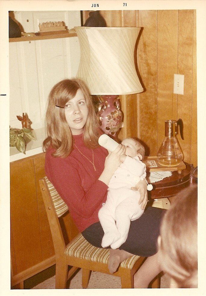 a woman sitting in a chair holding a baby next to a table with a lamp on it