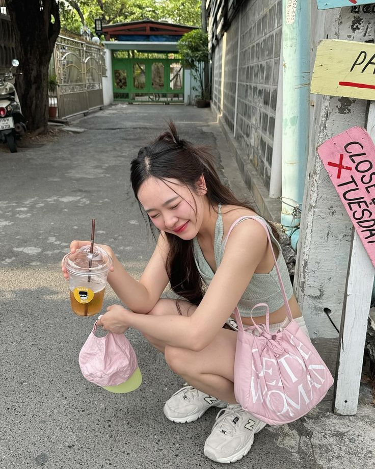 a woman sitting on the ground holding a pink bag and a drink in her hand