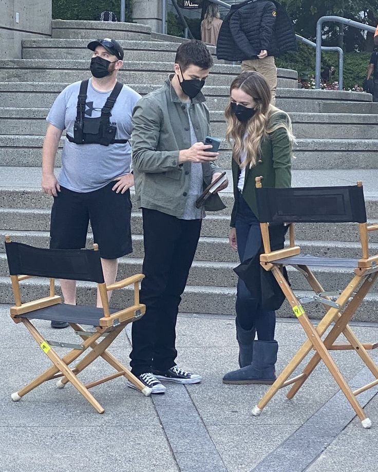 three people wearing masks standing in front of some stairs with chairs on each side and one person looking at his cell phone