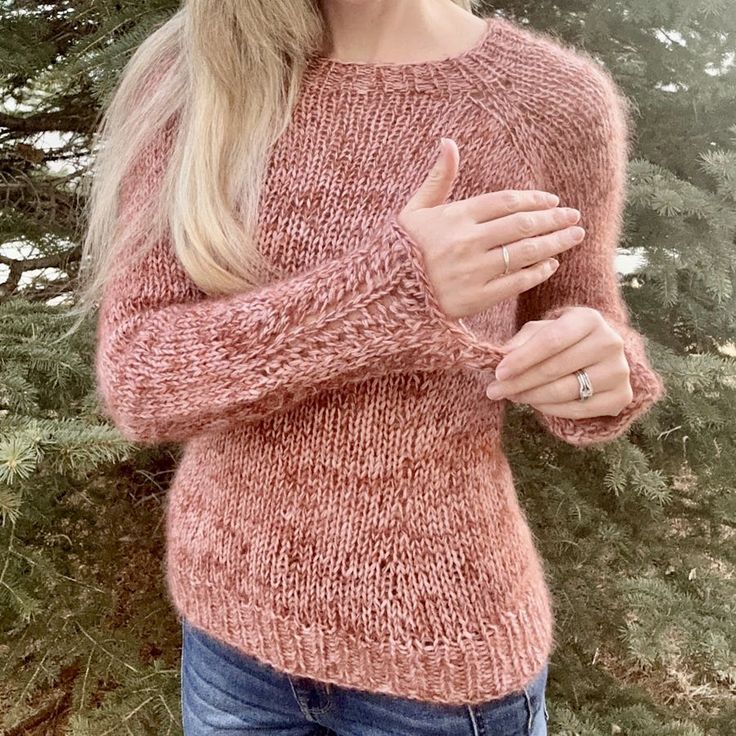 a woman wearing a pink sweater standing in front of a tree with her hand on her chest