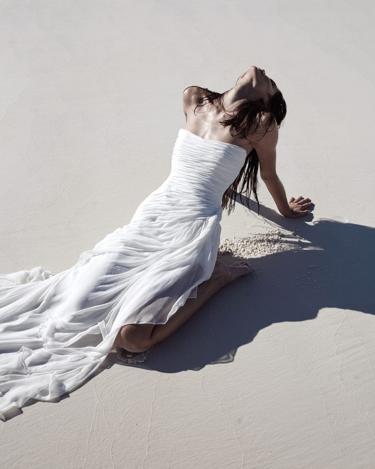 a woman in a white dress is laying on the sand with her hands behind her head