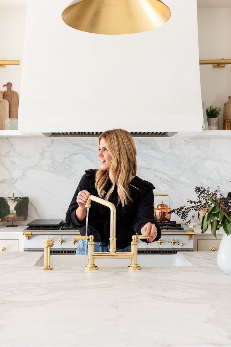 a woman standing in front of a kitchen sink with gold faucet and brass fixtures