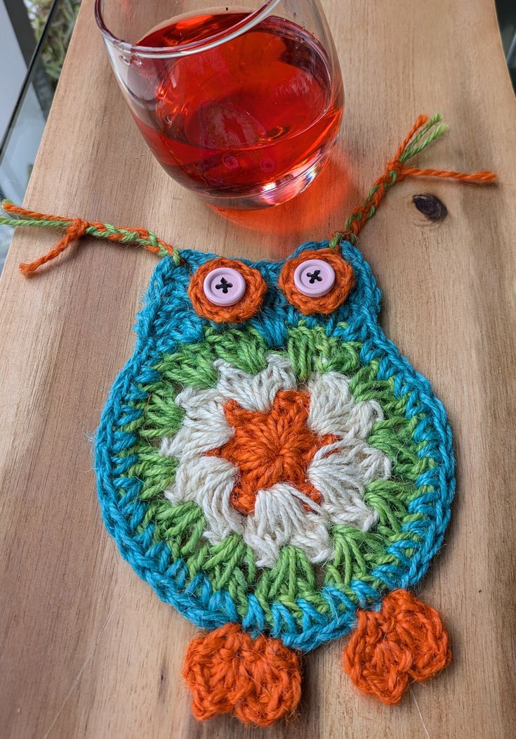 an owl shaped crochet coaster next to a glass of wine on a wooden table