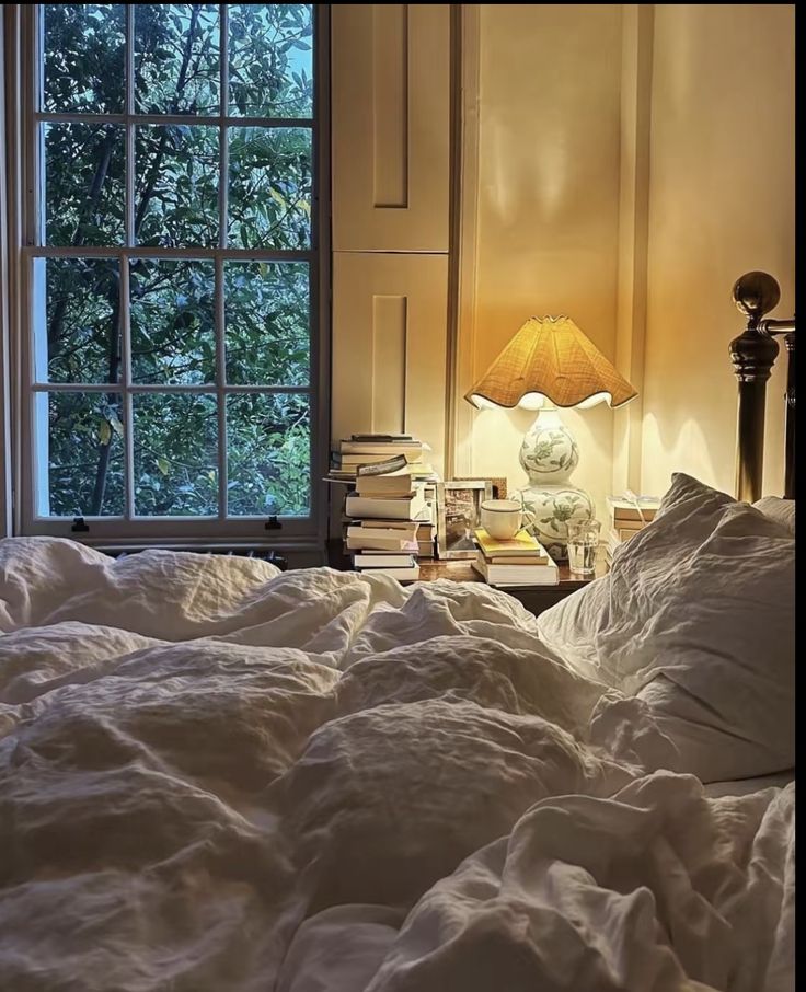 an unmade bed in front of a window with a lamp on the side table